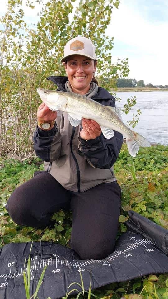 Een persoon knielt op een mat naast een rivier en toont trots zijn vangsten, terwijl een bont palet van bomen de serene achtergrond schildert.