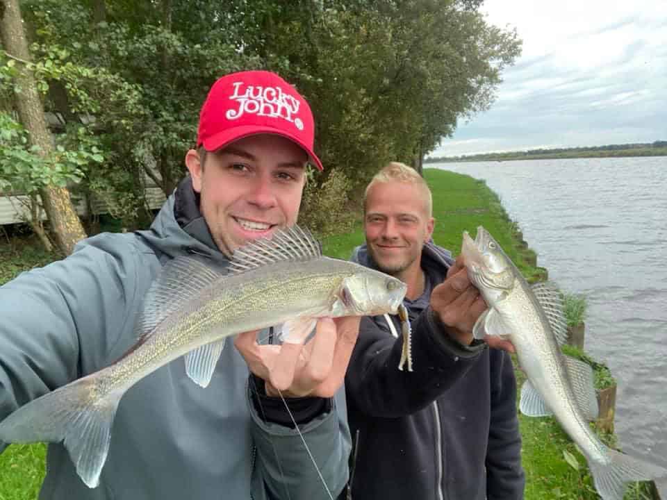 Twee mensen houden trots hun vangsten van de week vast aan een schilderachtig meer. Een van hen draagt een rode pet met "Lucky John" erop, wat flair toevoegt aan het gemengde palet van bomen en glinsterend water op de achtergrond.