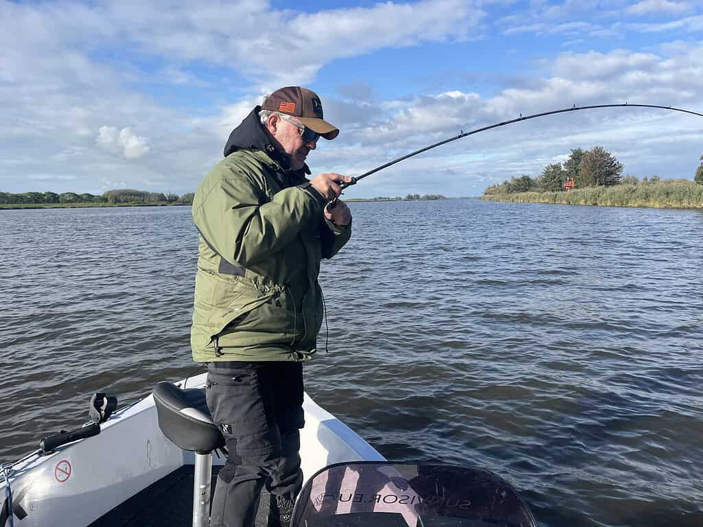 Een persoon in een groen jasje en met een pet op vist vanuit een boot op een groot wateroppervlak onder een bewolkte lucht.