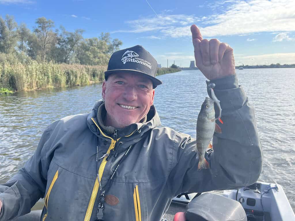 Lachende persoon in een jas en pet die een kleine vis aan een lijn vasthoudt terwijl hij op een boot in een meer staat, met bomen en een gebouw op de achtergrond en een gedeeltelijk bewolkte lucht.