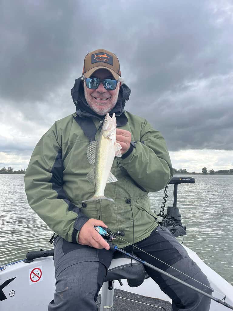 Een persoon zit in een boot, draagt een pet en een zonnebril, en houdt een vis vast met een hengel in zijn hand, tegen een bewolkte achtergrond.