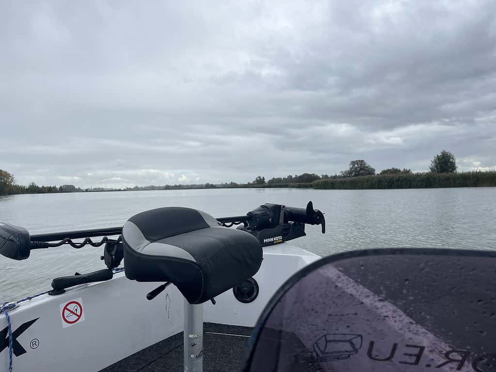 Uitzicht vanaf een boot op een kalme rivier onder een bewolkte hemel, met een zwart-witte stoel en een motor zichtbaar op de voorgrond.