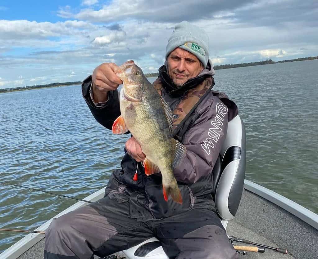 Zittend in een boot op het meer, met hoed en jas aan, laat de visser trots zijn vangst van de week zien: een grote vis die extra spanning toevoegt aan deze bomvolle editie.