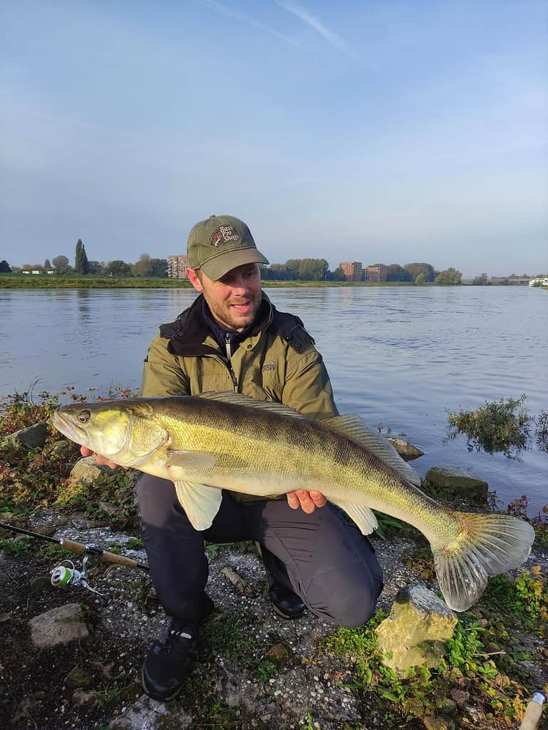 Een persoon houdt trots een grote vis vast bij de rivier, met een pet en buitenkleding. De scène legt de essentie van "vangsten van de week" vast, met glinsterend water onder een heldere hemel en verre gebouwen die bijdragen aan de pittoreske achtergrond.