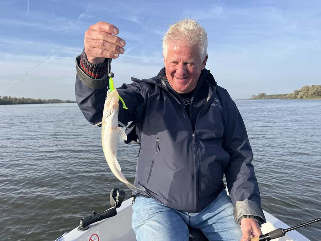 Een oudere man in een boot houdt een kleine vis omhoog die hij aan een vislijn heeft gevangen en kijkt er lachend naar.