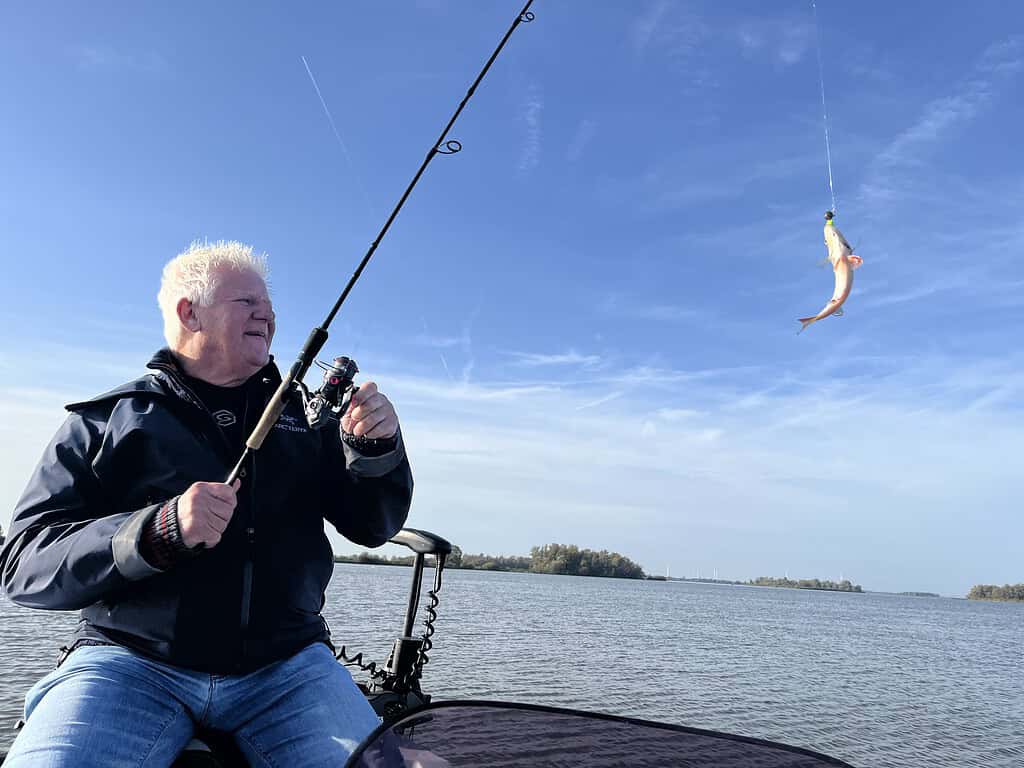 Een persoon op een boot houdt een hengel vast en haalt een kleine vis binnen boven kalm water onder een helderblauwe lucht.