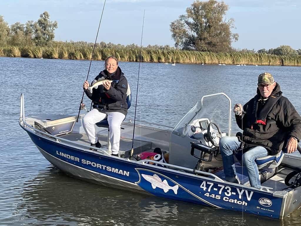 Twee mensen vissen op een blauwe boot met het label "Linder Sportsman 447 Catch" in een kalm waterlichaam, omringd door hoog gras en bomen. Een van hen houdt een hengel vast; beiden dragen jassen.