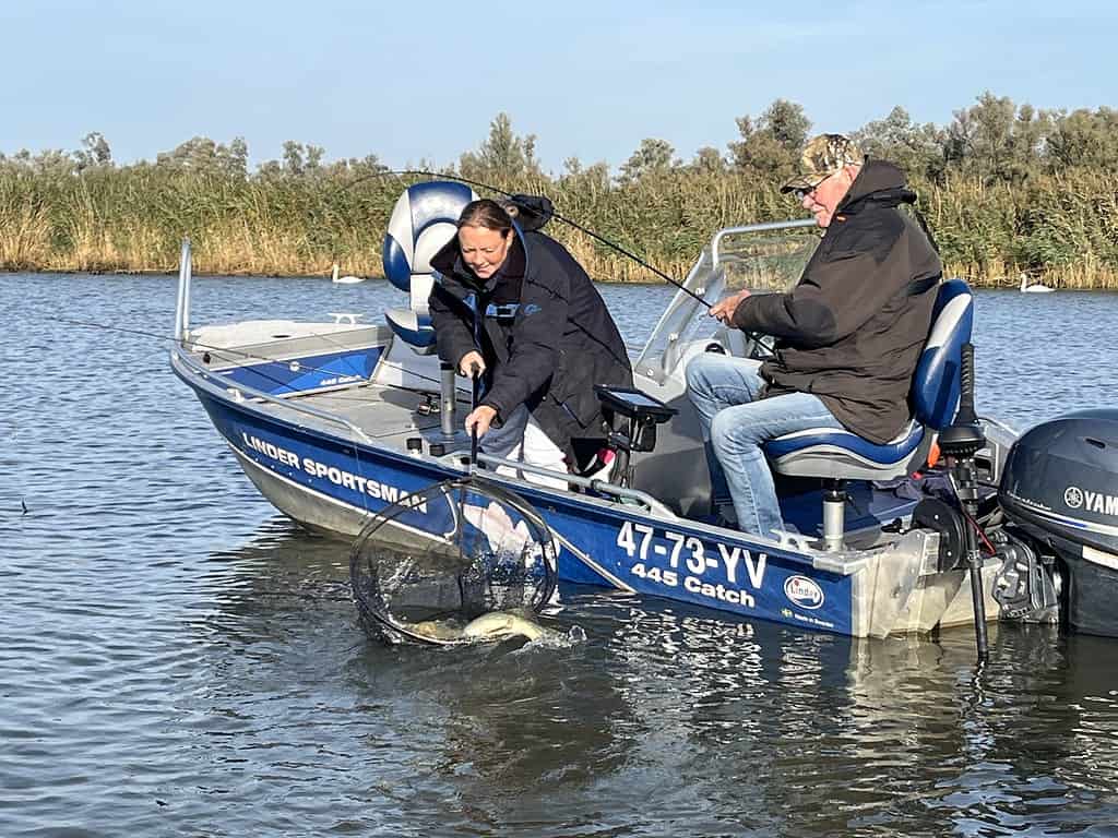 Twee mensen in een kleine vissersboot op een meer, die een grote vis binnenhalen met een net. Eén persoon staat, de ander zit. Omringend riet en een heldere lucht maken het tafereel compleet.