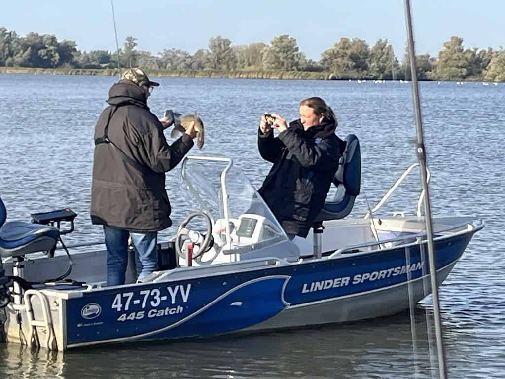 Twee mensen op een kleine boot; de een houdt een vis vast terwijl de ander een foto maakt. De boot ligt op een meer met bomen op de achtergrond.