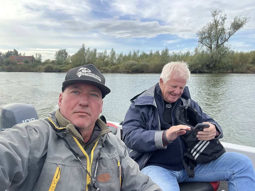 Twee mannen in jassen zitten in een boot op een rivier. De ene man neemt een selfie, terwijl de andere op zijn telefoon kijkt. Bomen langs de oever van de rivier op de achtergrond.