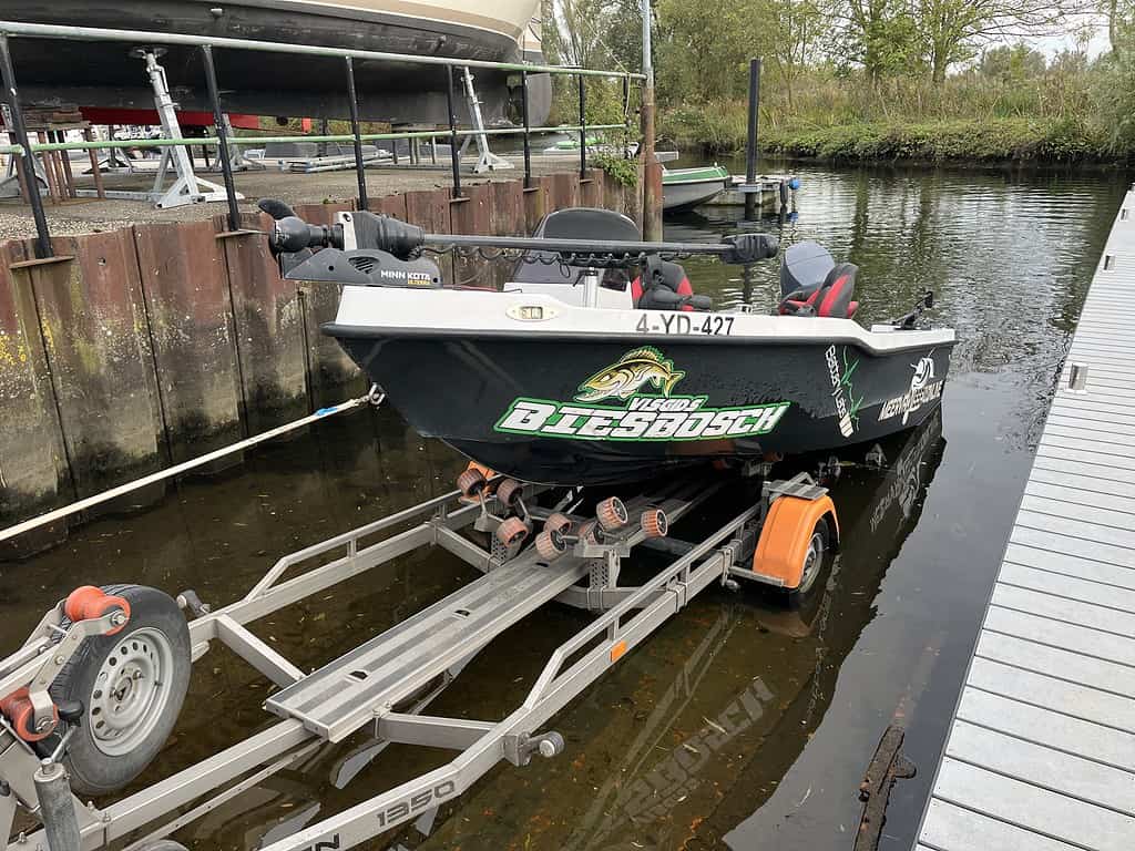 Een kleine vissersboot met sponsorstickers ligt op een trailer naast een steiger in een jachthaven.