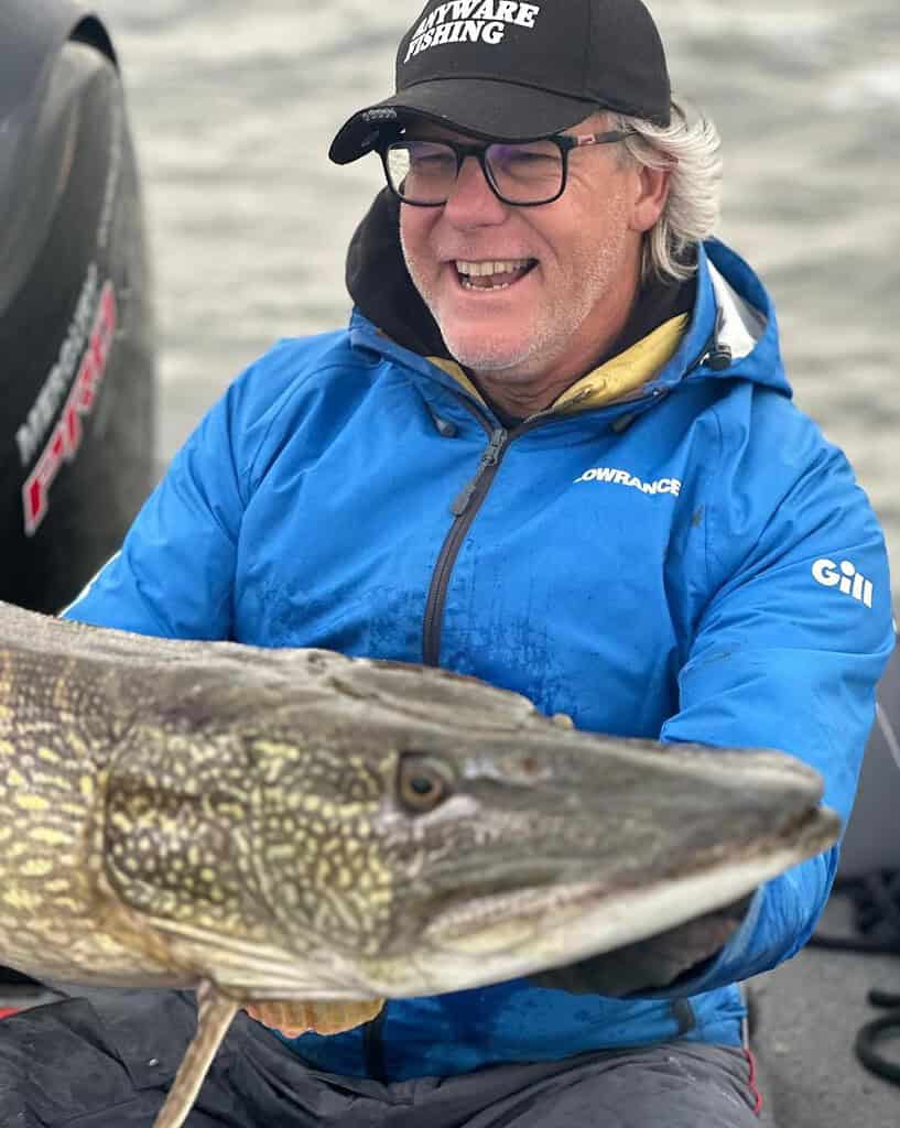 Een persoon in een blauw jasje en met een pet lacht terwijl hij een grote snoek vasthoudt op een boot, genietend van een perfecte dag vissen op het Volkerak.