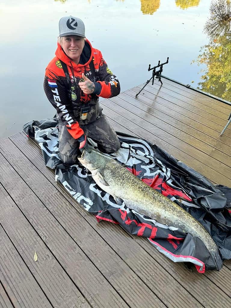 Een persoon knielt op een steiger en toont trots de vangsten van de week, een prachtige vangst, op een mat bij het water.