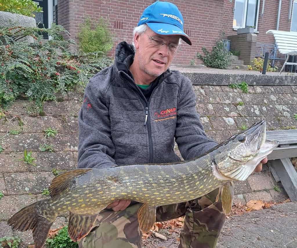 Een persoon met een pet en jas toont trots zijn vangsten, een grote vis, terwijl hij buiten zit, met een bakstenen bouwwerk op de achtergrond, en een moment vastlegt van een avontuurlijke week.