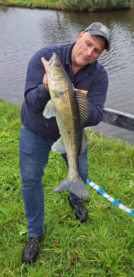 Een persoon met een pet op toont trots zijn vangst aan de oever van de rivier. Hij staat op de grasgrond en viert een van de vele succesvolle vangsten.