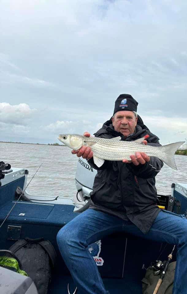 Een oudere man zit in een boot op het water en houdt zijn vangst van de week, een vis, met beide handen vast. Hij draagt een zwarte jas en muts onder de bewolkte lucht, waarmee hij zijn triomf te midden van de uitdagingen van de natuur laat zien.