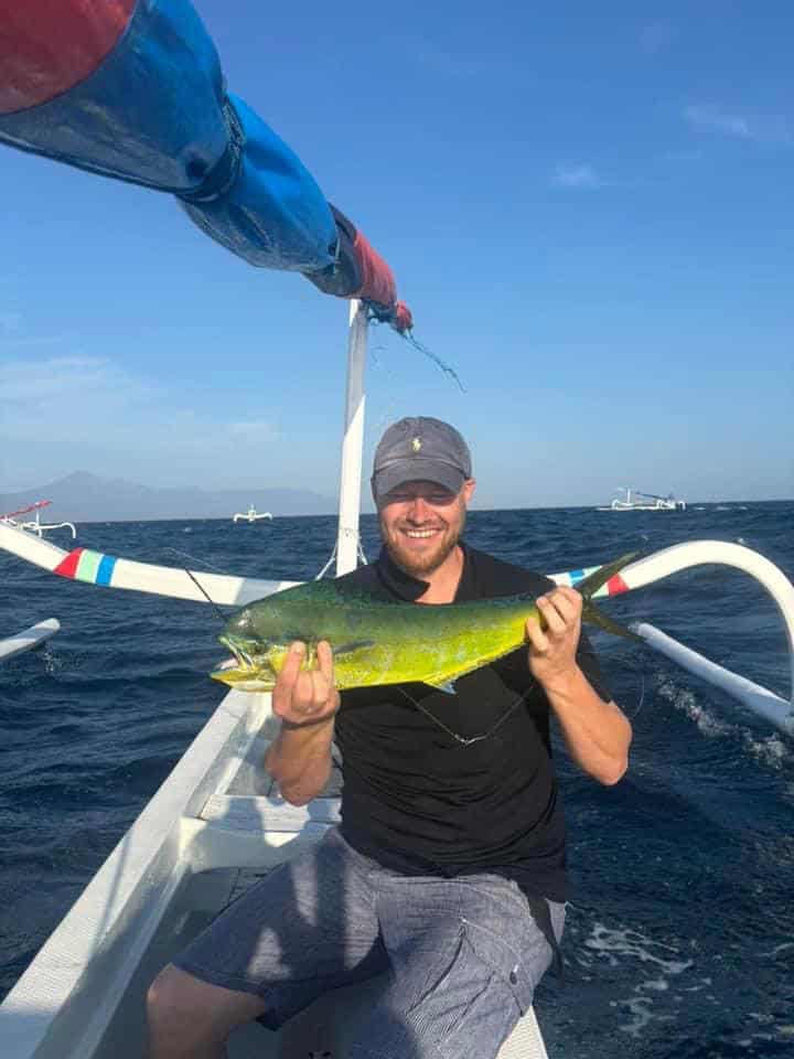 Man zit in een bootje op het water, houdt een geelgroene vis in beide handen vast en lacht trots naar zijn vangsten van de week.