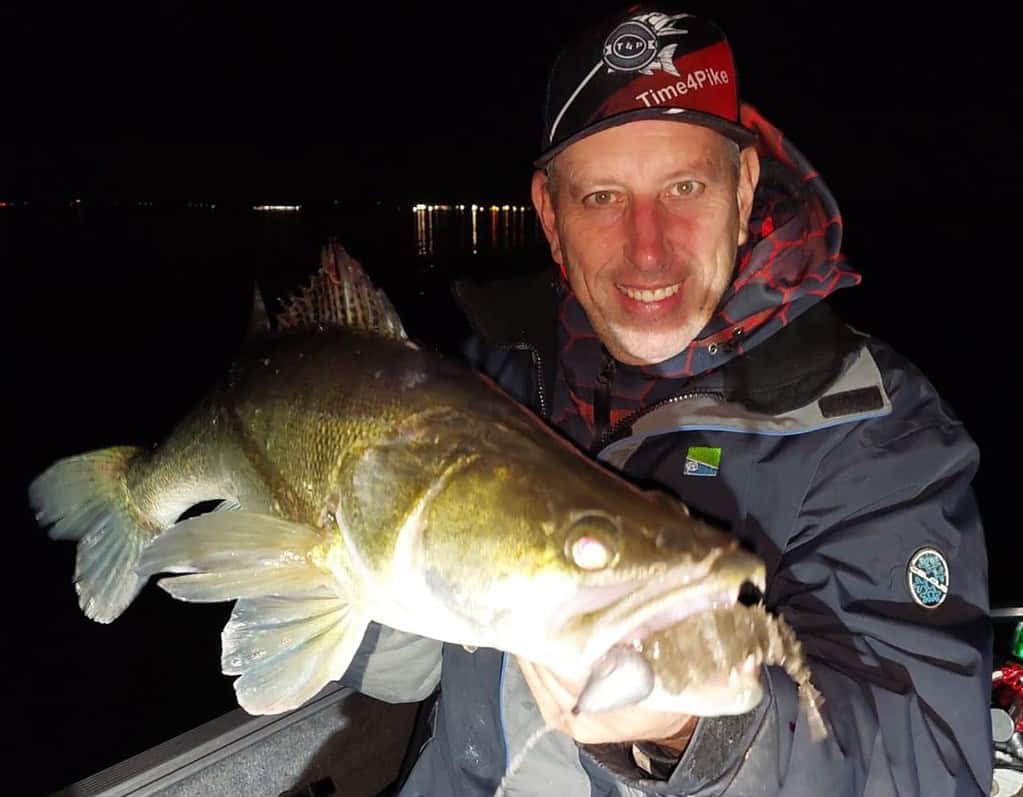 Een persoon houdt's nachts trots een grote vis vast op een boot, lachend naar de camera. Ze dragen een jas en een pet met "Time4Pike" erop, en vieren hun vangsten tegen een donkere achtergrond met lichtjes in de verte.