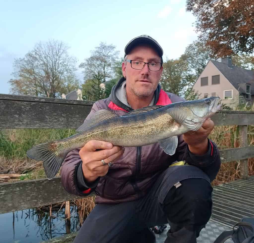 Een man toont trots zijn vangsten terwijl hij op een houten brug staat, met bomen en huizen op de achtergrond.