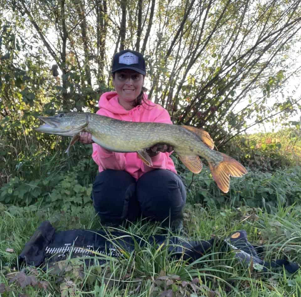 Een persoon in een roze hoodie en pet knielt op het gras en toont trots zijn vangst van de week: een grote vis. Struiken en bomen vormen een weelderige achtergrond.