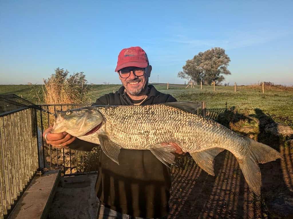 Een persoon met een rode pet en bril houdt trots een grote vis vast, wat een triomfantelijke week van vangsten in een buitenomgeving markeert. De heldere lucht en weelderige vegetatie vormen een pittoresk decor voor deze indrukwekkende vangst.