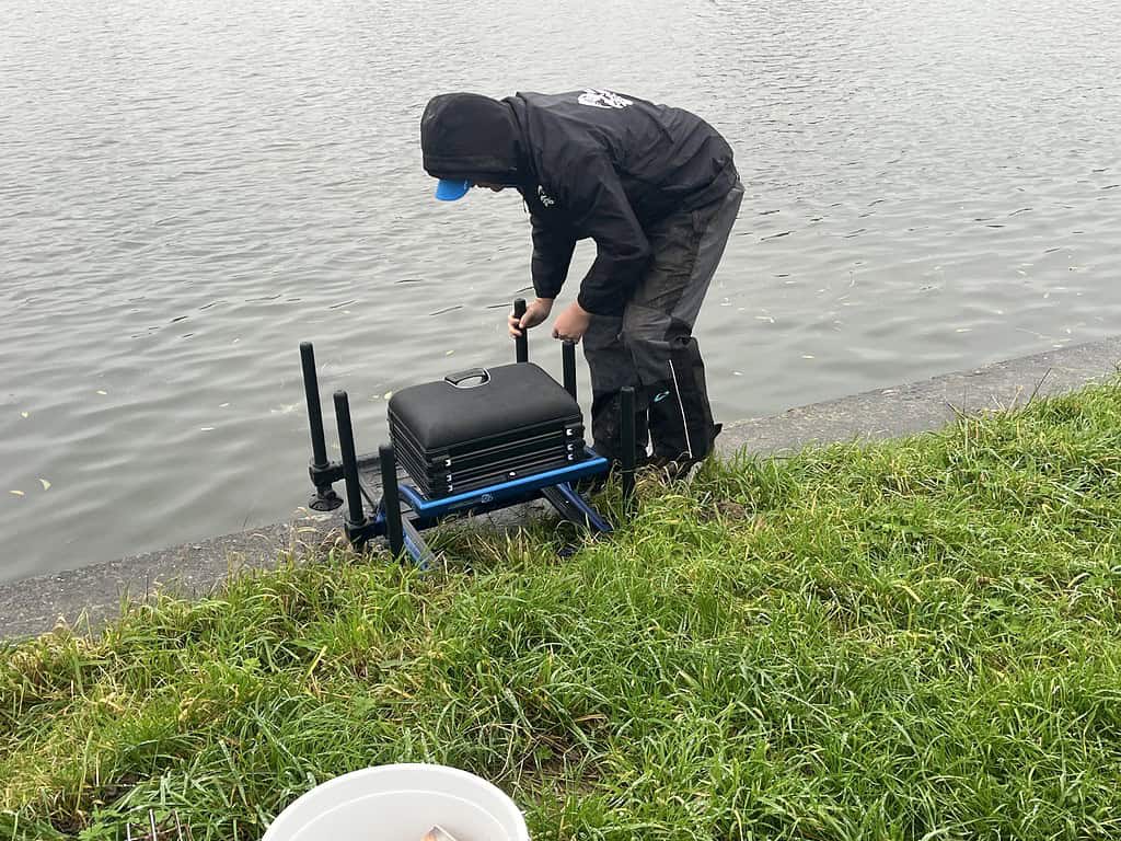 Jelle Zwijnenburg is druk bezig met het uitrusten van zijn visgerei op het grasveld bij een rustig kanaal, ter voorbereiding op een heerlijk dagje feedervissen.