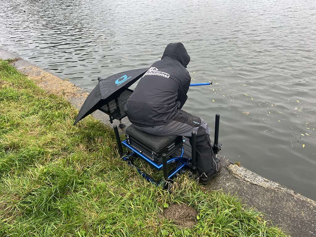 Persoon zittend op een visstoel aan het kanaal, gekleed in outdoorkleding met een paraplu eraan, met het gezicht naar het water. Feedervissen technieken oefenen die doen denken aan Jelle Zwijnenburg's advies om de perfecte vis te vangen.