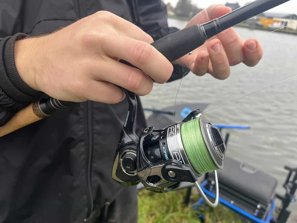 Jelle Zwijnenburg houdt behendig een hengel vast met een groene lijn terwijl hij aan het feedervissen is bij het rustige kanaal.