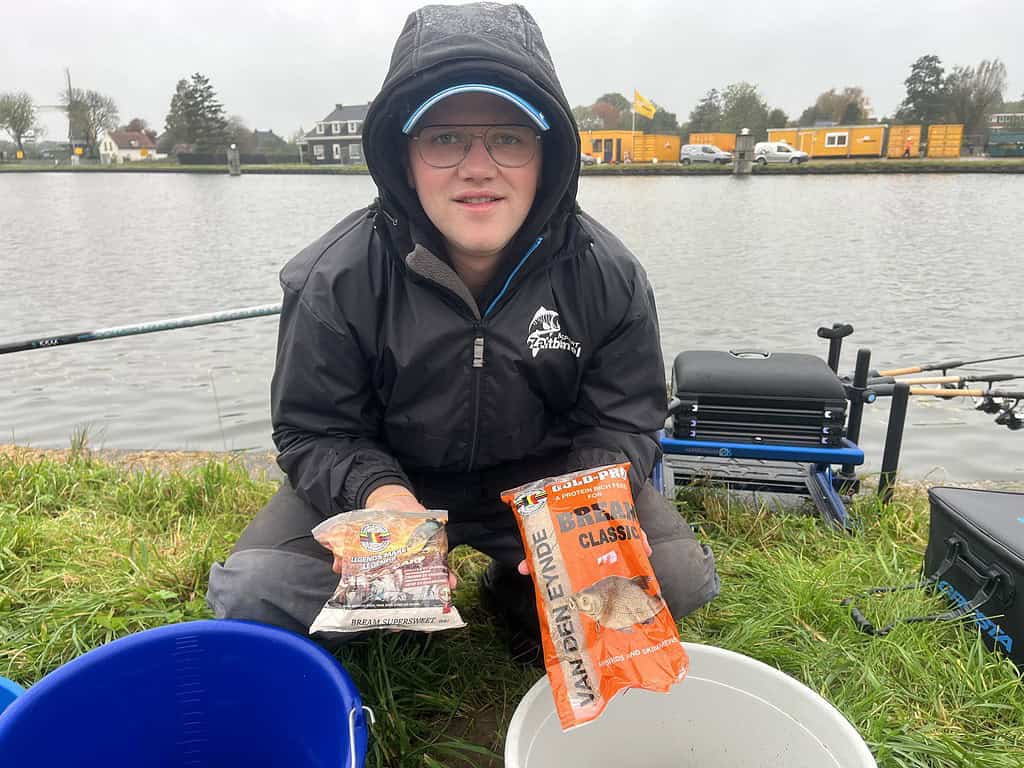 Een persoon gekleed in outdoorkleding houdt twee zakken met visvoer vast bij het kanaal, met feedervisuitrusting en schilderachtige huizen als pittoreske achtergrond.