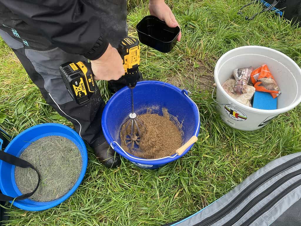 Een persoon gebruikt een boor om de inhoud van een blauwe emmer op het gras te mengen, met een andere emmer met items zoals brood in de buurt. Het lijkt erop dat ze zich voorbereiden op wat feedervissen bij het kanaal, geïnspireerd door de technieken van Jelle Zwijnenburg.