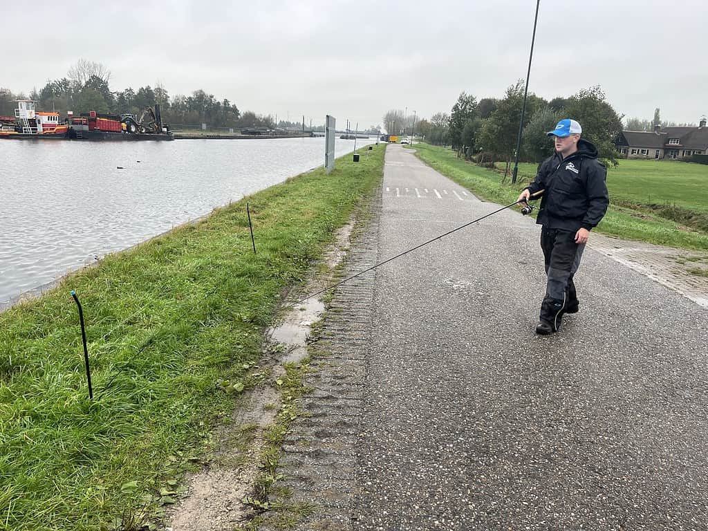 Een persoon wandelt langs een kanaalpad met een hengel in de hand voor een middagje feedervissen op een bewolkte dag.