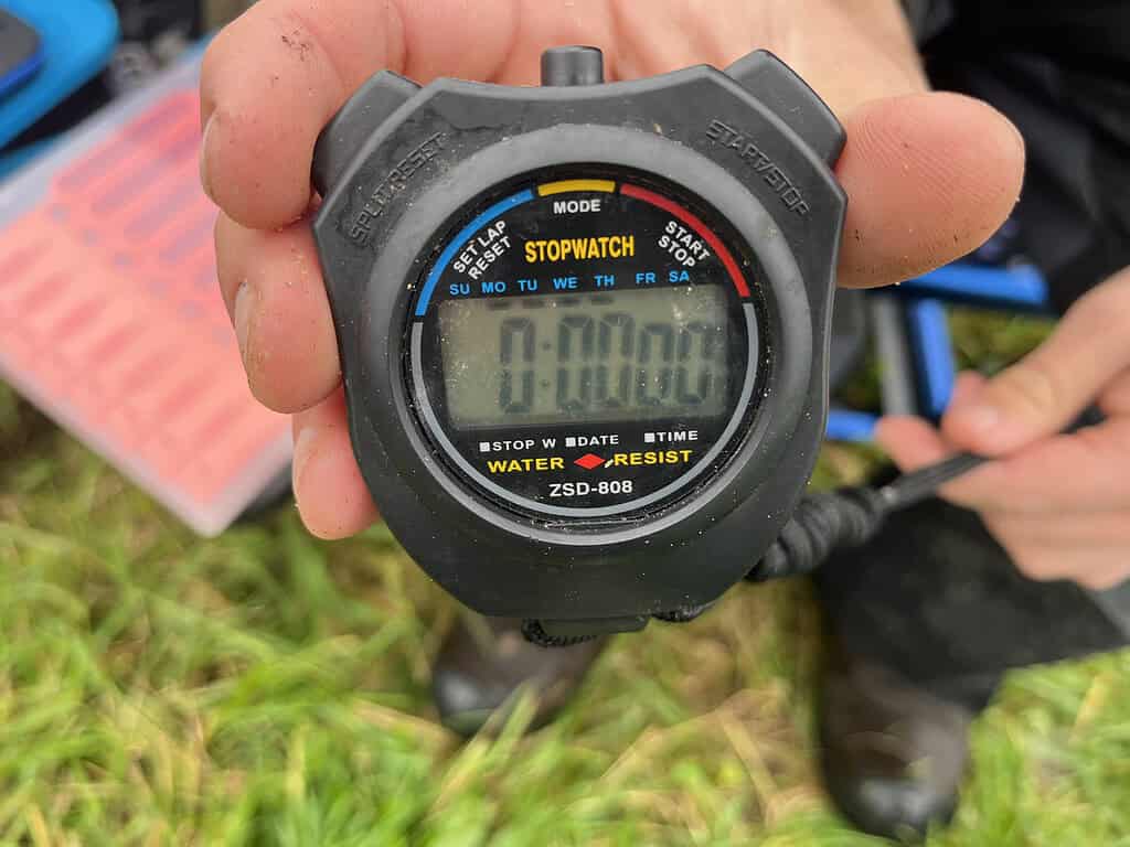 Een hand houdt een stopwatch vast die op 0:00:00 staat, klaar om de tijd te meten bij het kanaal. Gras is zichtbaar op de achtergrond, wat wijst op een dag feedervissen terwijl de expertise van Jelle Zwijnenburg wordt gekanaliseerd.