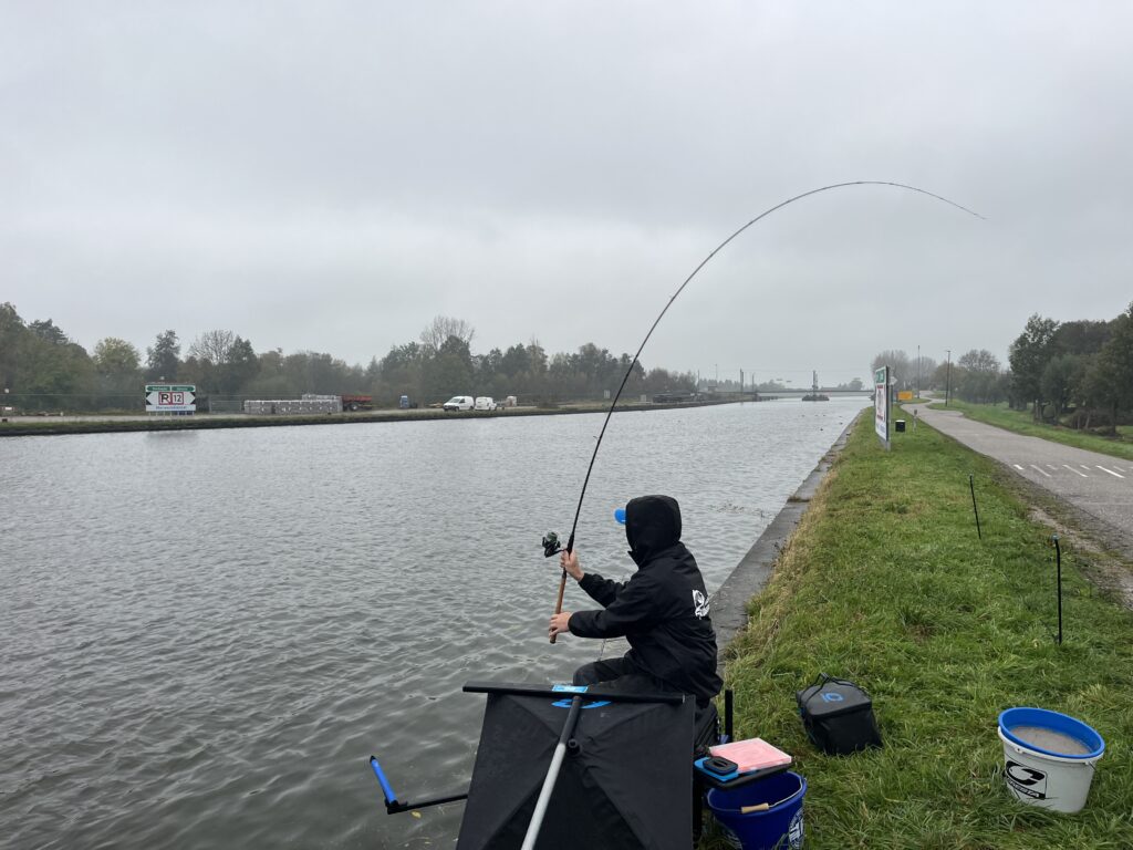 Jelle Zwijnenburg zit aan het kanaal onder een bewolkte hemel, verdiept in feedervissen. Omringd door zijn visgerei, vormen de weg en de bomen een vredig decor voor zijn middag aan het water.