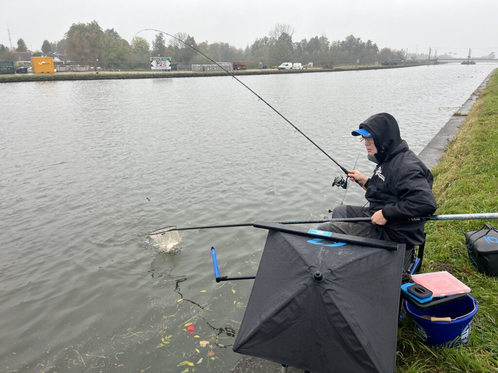 Jelle Zwijnenburg, gekleed in regenkleding, beoefent feedervissen langs het kanaal. Comfortabel zittend op een kruk, omringd door een paraplu en zijn vertrouwde visgerei.