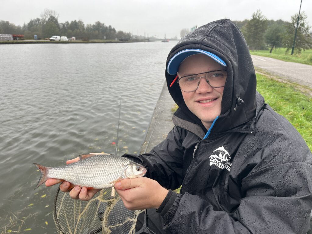 Jelle Zwijnenburg, gekleed in een regenjas, houdt op een bewolkte dag trots een vis vast aan de oever van de rivier. Het is een symbool van de serene charme van het vissen op het kanaal.