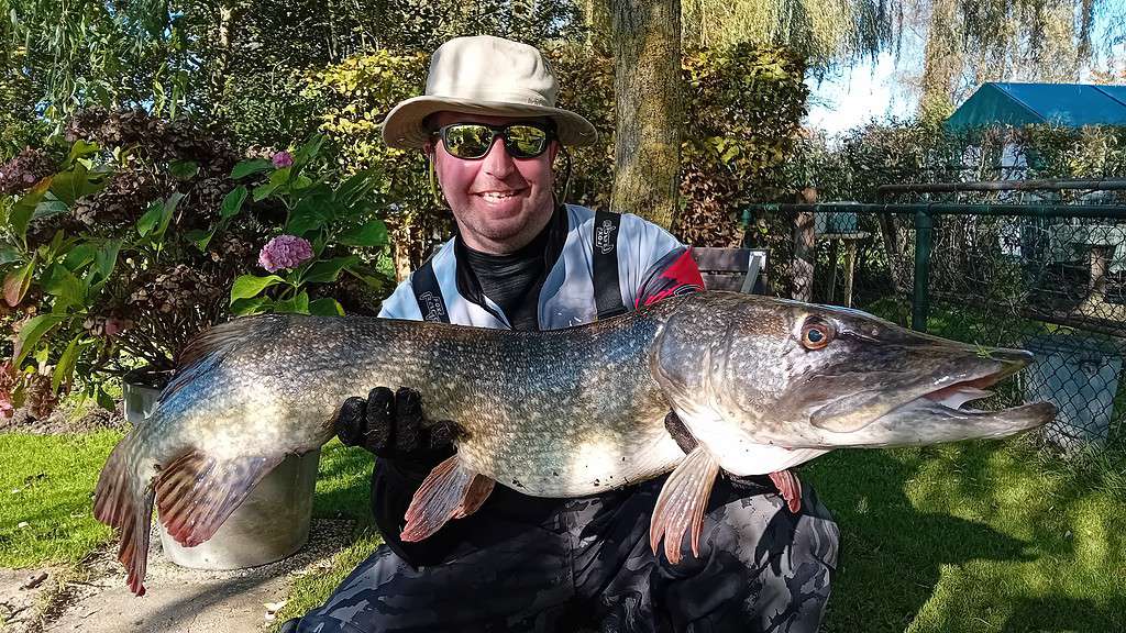 Een man met een hoed en zonnebril toont trots zijn week's vangsten, met een grote vis in zijn hand, mogelijk een snoek, buiten. Hij lacht in zijn outdoor-uitrusting, omringd door weelderig groen.