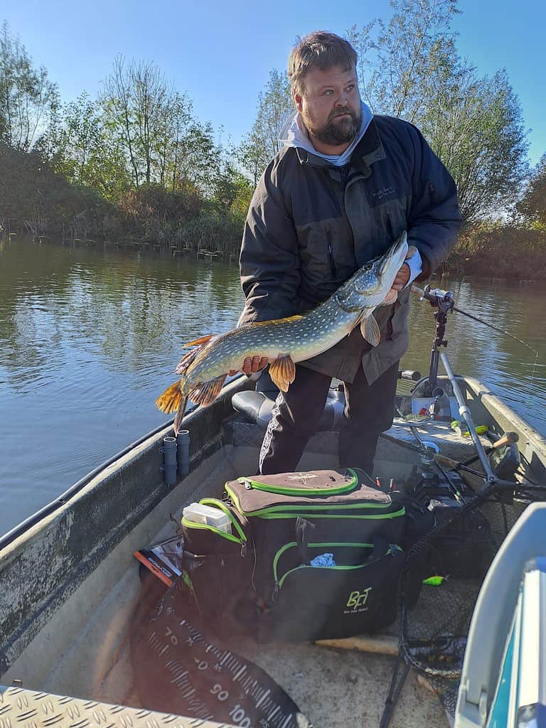 Een man staat op een boot, trots een grote vis vasthoudend, mogelijk een snoek, en toont zijn indrukwekkende vangsten boven een tacklebox. Bomen en water vormen de achtergrond onder de heldere lucht.