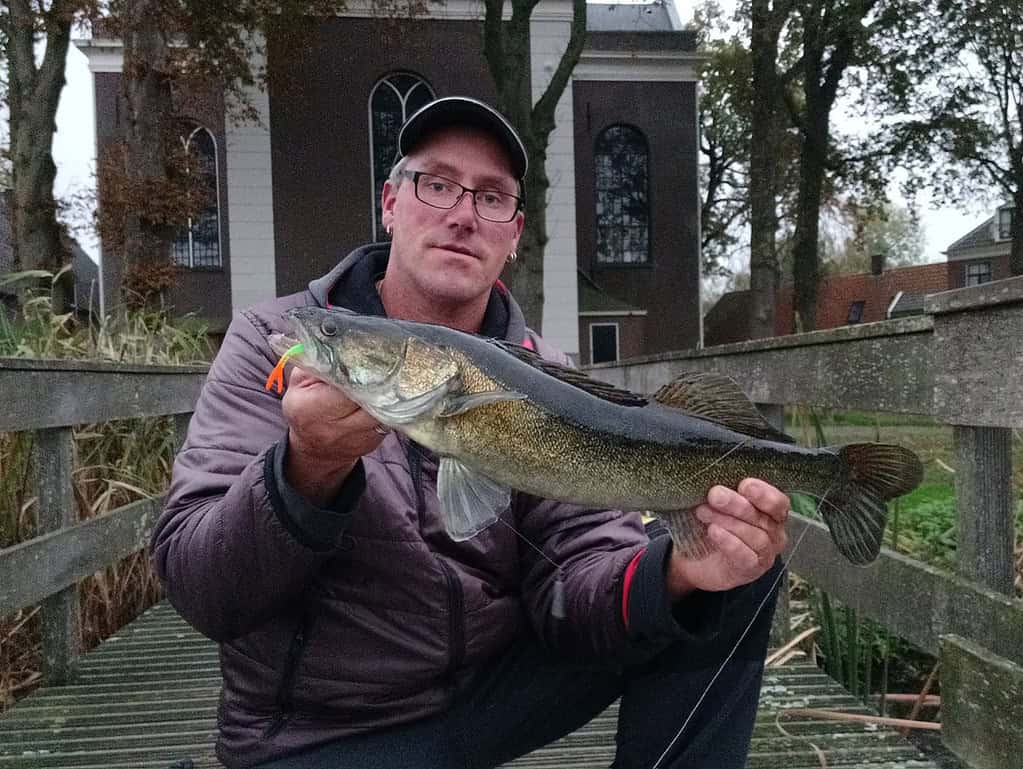 Een persoon die trots een grote vis vasthoudt op een houten brug, tandjes die glinsteren van plezier, terwijl een pittoresk kerkje en weelderige bomen het perfecte decor vormen voor dit opmerkelijke vangsten-moment.