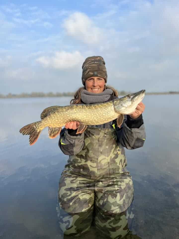 Onder een gedeeltelijk bewolkte lucht knielt een persoon in camouflagekleding in ondiep water, trots hun vangst van de week tonend - een grote vis die glinstert in de zon. Het is alsof "Jullie hebben het geflikt" door de scène galmt, dit indrukwekkende moment vierend.