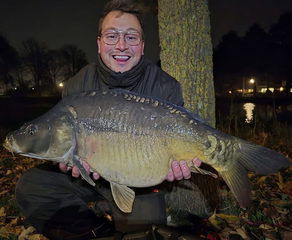 Een persoon lacht terwijl hij zijn indrukwekkende vangsten buiten vasthoudt in de nacht, staand naast een boom, met twinkelende lichtjes op de achtergrond. Ze hebben het echt geflikt deze week met zo'n grote vangst!