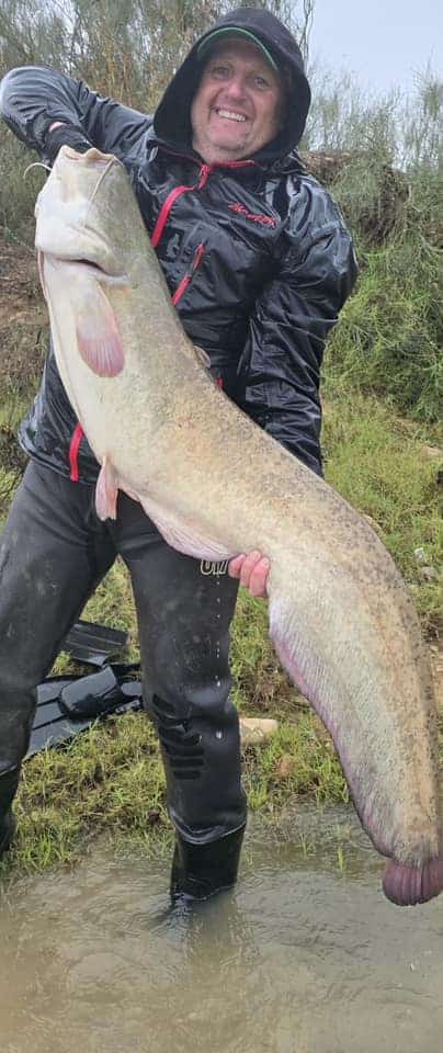 Een persoon in waterdichte kleding laat trots zijn vangsten, een grote vis, zien bij een grasachtig en nat gebied.