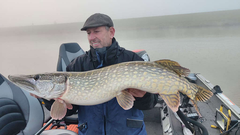 Een persoon op een boot houdt trots zijn nieuwste vangsten, een grote vis, vast tegen de mistige achtergrond van een mistig meer.