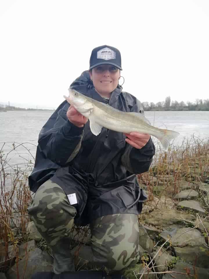 Een persoon in een camouflagebroek en een zwart jasje toont trots zijn vangsten aan de oever van de rivier, met een pet en een zonnebril op.