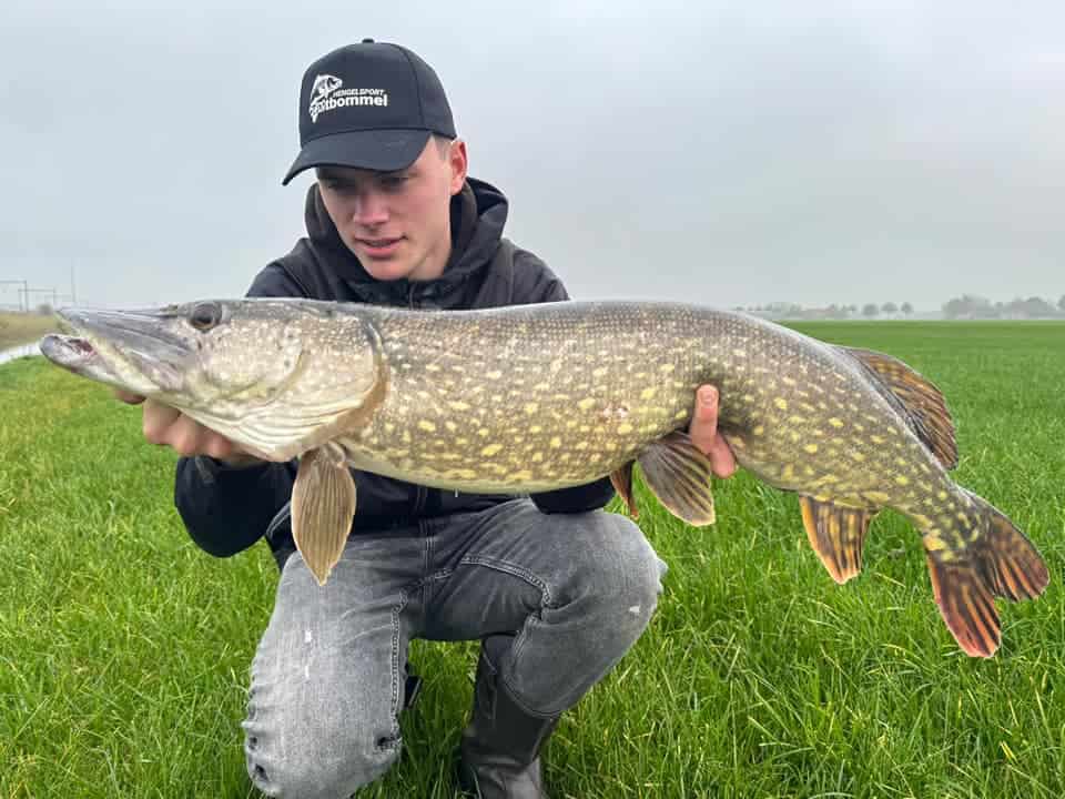 Een man knielt op het weelderige gras en toont trots zijn indrukwekkende vangsten: een grote vis die hij heeft gevangen ondanks de stevige wind die buiten waait.