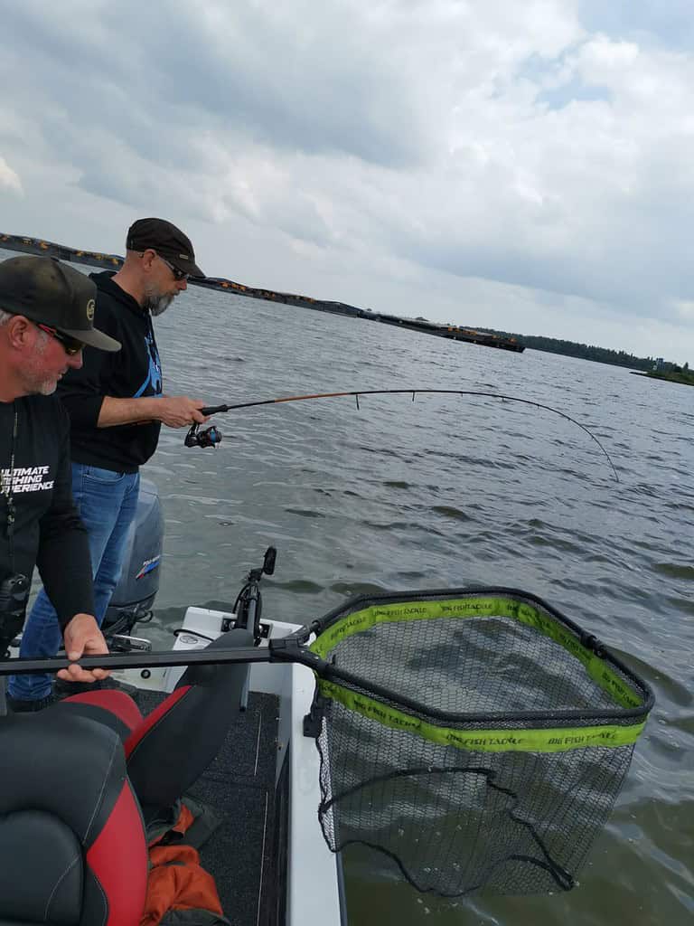 Twee mensen vissen op een boot, met hengels in de hand. Een persoon bereidt een net voor, in de hoop een visje voor de pan te vangen. Bewolkte lucht en kalm water op de achtergrond.