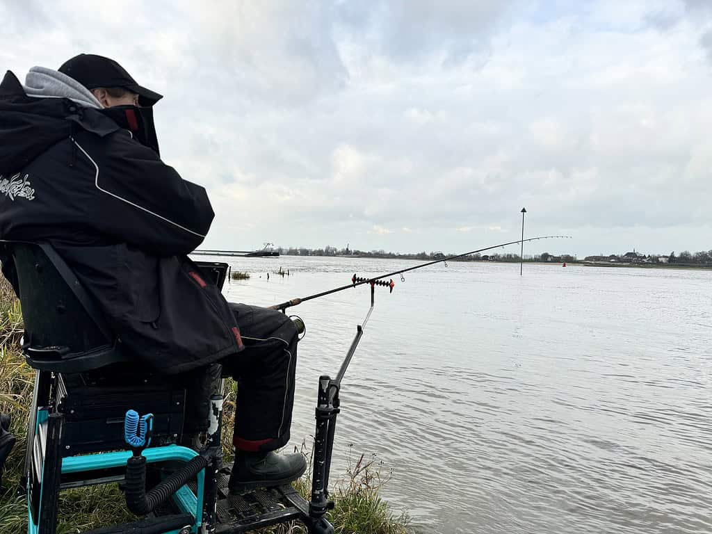 Een persoon zit aan de rivier de Waal en is op een bewolkte dag met een hengel aan het feedervissen terwijl het hoge water zachtjes tegen de oever klotst.