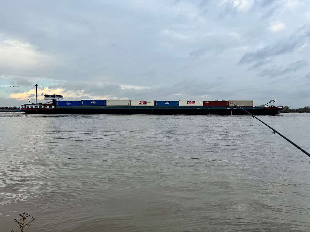 Een groot vrachtschip met meerdere gekleurde containers vaart op een bewolkte dag over de Waal. Een hengel, perfect voor feedervissen, is zichtbaar op de voorgrond aan de waterkant, wat wijst op mogelijke avonturen bij hoog water.