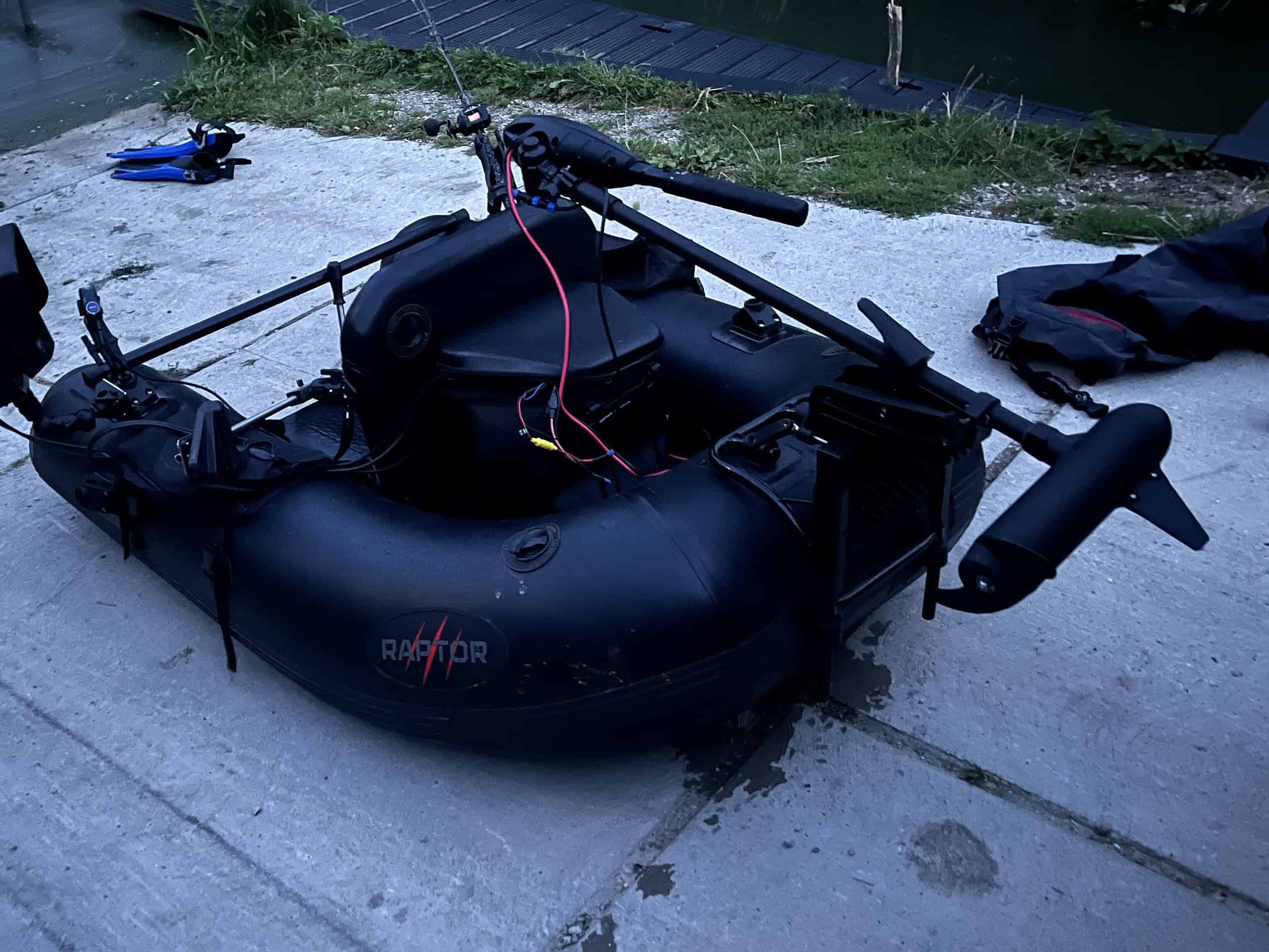 Een kleine zwarte opblaasbare boot met een gemonteerde motor en stuur op een betonnen ondergrond in de buurt van water.