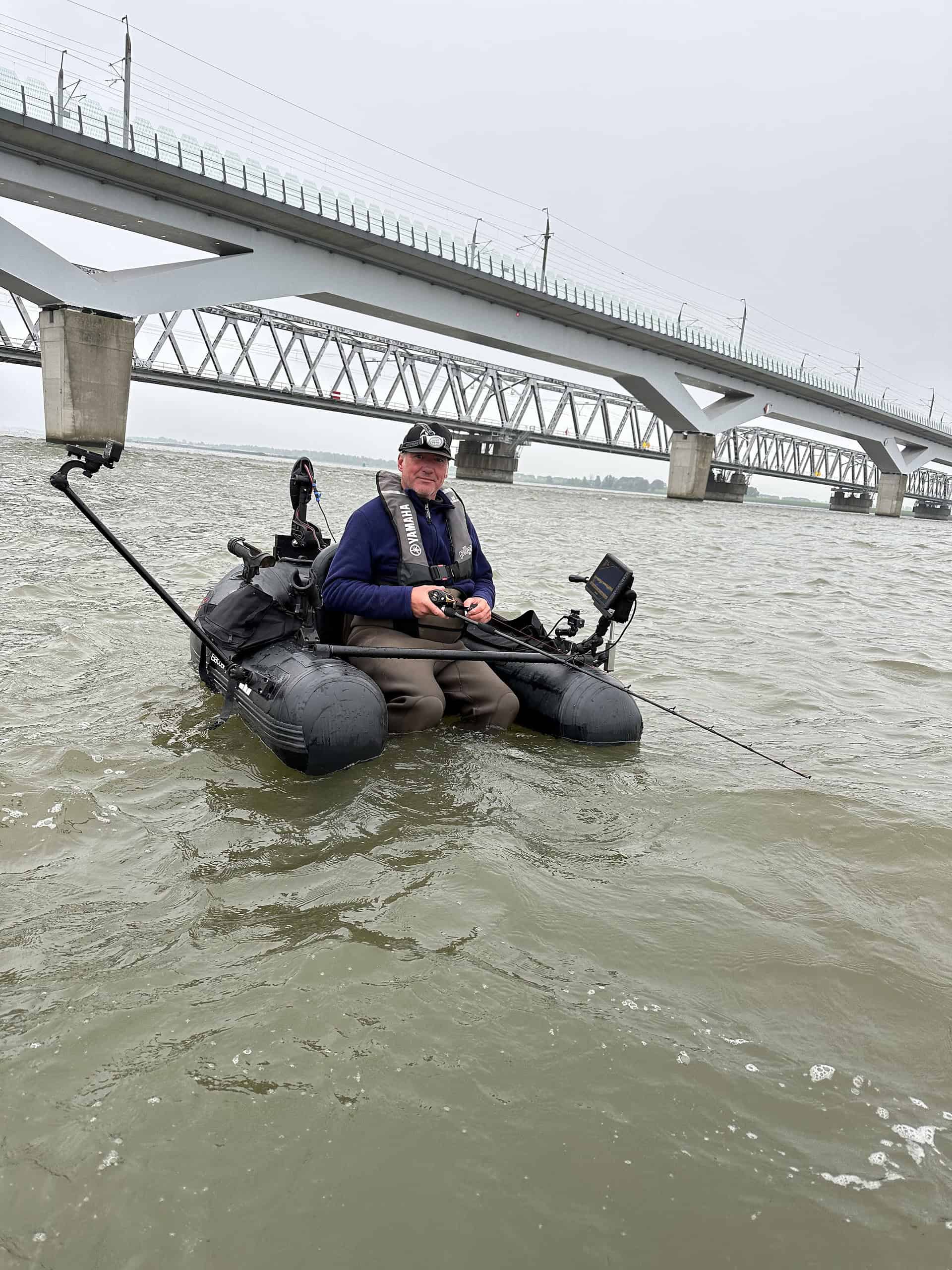 Een persoon vist in een kleine, zwarte opblaasboot op een rivier, met een brug op de achtergrond.