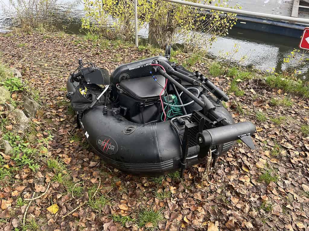 Een opblaasboot ligt op de met bladeren bedekte grond bij het water, uitgerust met visgerei en een elektromotor, klaar om een spannende week af te sluiten.